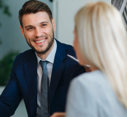 A man in a blue suit smiles while discussing home loans with a woman with blonde hair. Both are engaged in conversation in an indoor setting.